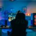 Rückenaufnahme eines Spielers, der ein First-Person-Shooter-Online-Videospiel auf seinem leistungsstarken Computer spielt. Zimmer und PC haben kalte bunte Neon-LED-Lichter. Gemütlicher Abend zu Hause.