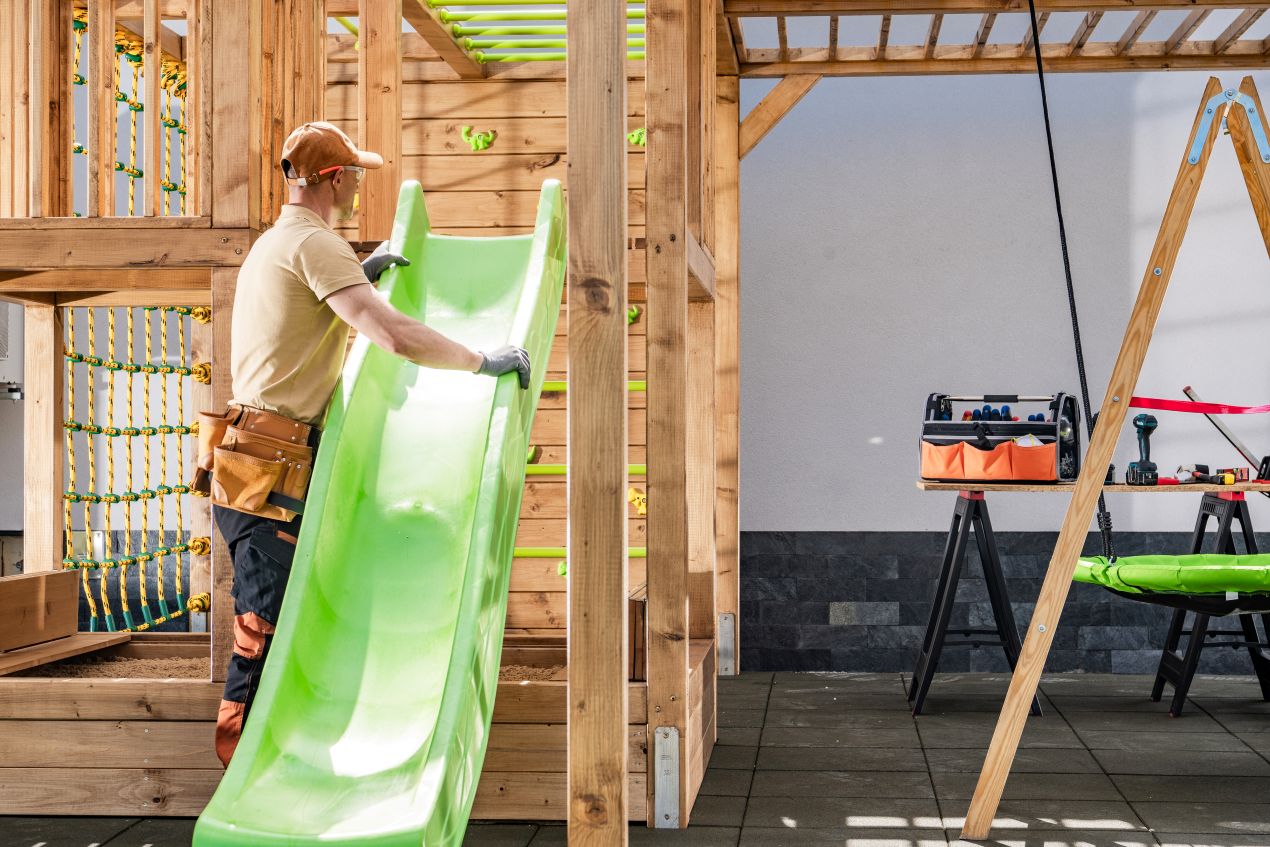Handwerker montiert gruenes Spielplatzgeraet auf einem Holzspielplatz fuer Kinder mit Fokus auf Sicherheit und Langlebigkeit